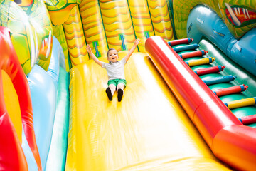 Little boy child rides on an inflatable multi-colored slide. - obrazy, fototapety, plakaty