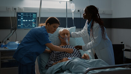 Patient with breathing problems fainting in hospital ward bed at modern equipment medical facility. Nurse and african american doctor helping old man with stethoscope and oxygen tube