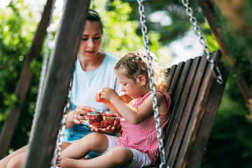 A happy mother is sitting on a swing with her charming daughter, who is eating fresh strawberries. Childhood, motherhood, parenthood. Time together, summer time