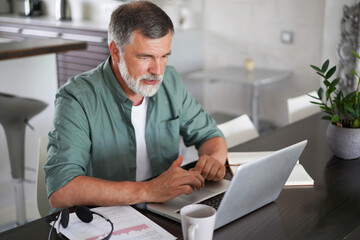 Handsome mature man in casual suit sitting at the table in home office and working at laptop