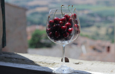 glass, wine goblet filled with lots of cherries standing on a windowsill against a blurred tuscany background 