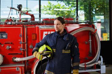 Portrait of male firefighter in protective gear holding helmet while standing beside fire truck,...