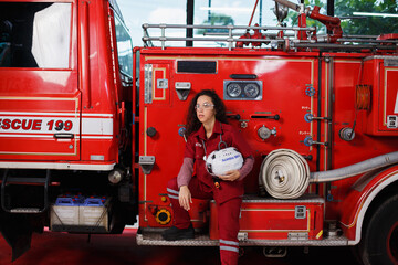 Female firefighter in red uniform holding helmet while resting beside fire truck, symbolizing strength, dedication, and resilience in emergency rescue service profession.