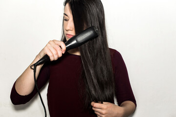 Beautiful caucasian young woman straightening her long black shiny hair using a flat iron isolated in a white background
