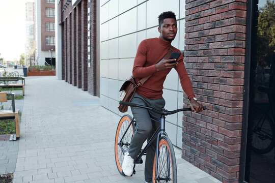 Young businessman using smartphone while riding bicycle along modern architecture