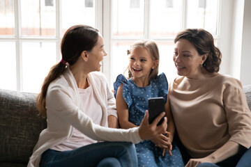 Excited three generations of Caucasian women sit on sofa at home have pleasant webcam virtual event on cellphone. Smiling girl with mother and grandmother talk speak on video call on smartphone.
