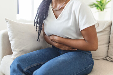 Fototapeta premium African American Woman in painful expression holding hands against belly suffering menstrual period pain, lying sad on home bed, having tummy cramp in female health concept