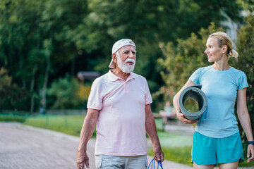 Active senior man and woman physiotherapist carrying exercise mats after sports workout in nature. Old man and his adult daughter holding yoga mats, walking along park path together, talking.