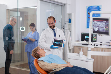 Stomatolog holding x-ray of senior woman sitting on orange chair in dentist cabinet. Medical teeth care taker holding patient radiography on tablet pc near patient standing up.