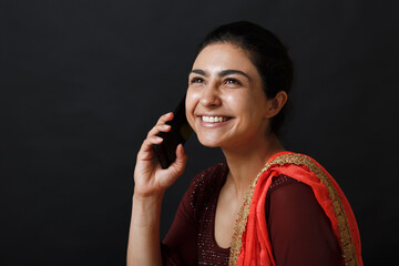 Portrait of smiling young adult indian woman in sari talking mobile phone against black background.