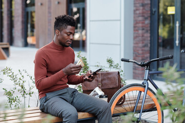 Obraz premium Serious African white collar worker eating sandwich and using smartphone