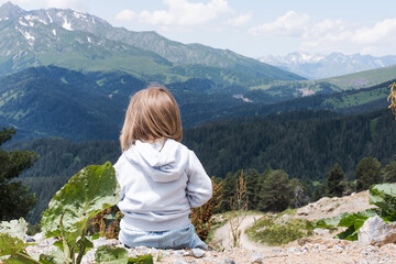 Naklejka na ściany i meble The child is sitting and looking at the landscape of the Caucasus Mountains. Outdoor travel, local travel.