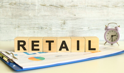 Wooden cubes lie on a folder with financial charts on a gray background. The cubes make up the word RETAIL.