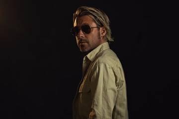 Man with blond hair and a stubble beard and dark aviator sunglasses wears a safari shirt. Looking over his shoulder.