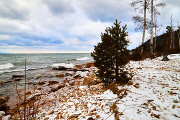Nature landscape with dramatic sky and clouds, lake with ice, shore with snow and dark trees in...