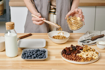 Hands of female putting muesli into bowl while preparing breakfast
