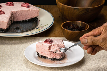 Male senior hand using metal spoon taking a bite from delicious homemade frozen strawberry cheesecake in white ceramic dish.