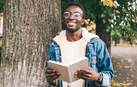 Portrait of young smiling african man student with a book looking away wearing an eyeglasses in autumn city park