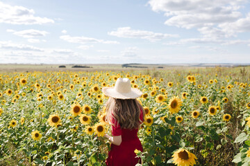 Beautiful young woman in red dress and a straw hat is standing against a yellow field of sunflowers. Summer time, cottagecore concept. Back view - Powered by Adobe