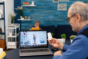 Senior man showing doctor pills bottle at webcam during video call. Elderly man discussing with healthcare practitioner in the course of remote call and wife is reading a book on sofa.
