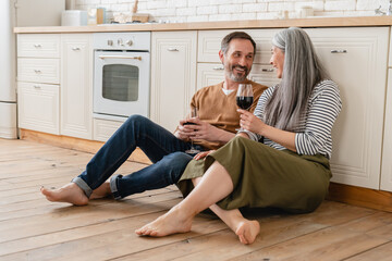 Mature family couple middle-aged wife and husband drinking wine in the kitchen, celebrating...