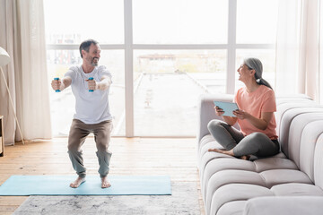 Mature husband doing sit-ups with dumbbells while his middle-aged wife watching fitness tutorial on tablet , avoiding physical exercises, being lazy at home on lockdown