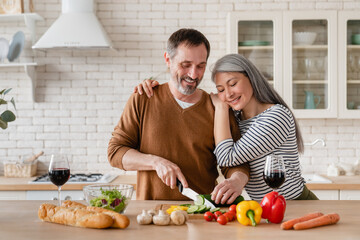 Happy middle-aged family cooking vegetable salad in the kitchen at home together. Mature wife...