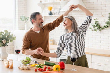 Happy active mature middle-aged couple family wife and husband dancing together while cooking vegetable salad, preparing vegetarian food meal in the kitchen at home. Love and relationship concept
