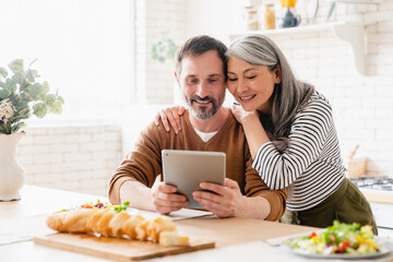 Mature middle-aged family couple wife and husband parents using digital tablet while having...