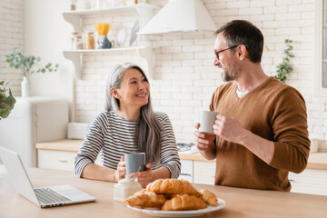 Obraz na płótnie Canvas Happy mature middle-aged couple family wife and husband having breakfast together in the kitchen, watching movies on laptop, e-learning, working from home. Love and relationship concept