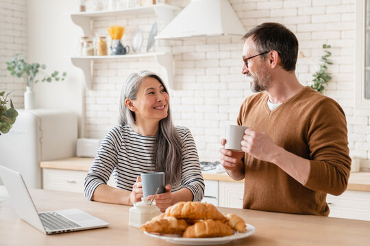 Happy mature middle-aged couple family wife and husband having breakfast together in the kitchen, watching movies on laptop, e-learning, working from home. Love and relationship concept