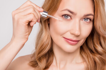 Portrait of attractive woman applying serum under eye freshness uplift effect isolated over grey pastel color background