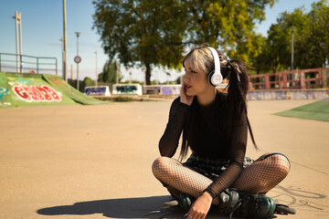 Cute young girl with punk style and with white headphones and inline skates sitting on the floor...