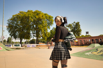 Young girl with pigtails, punk style, white headphones and inline skates hanging from the shoulder on their backs looking at the camera smiling.