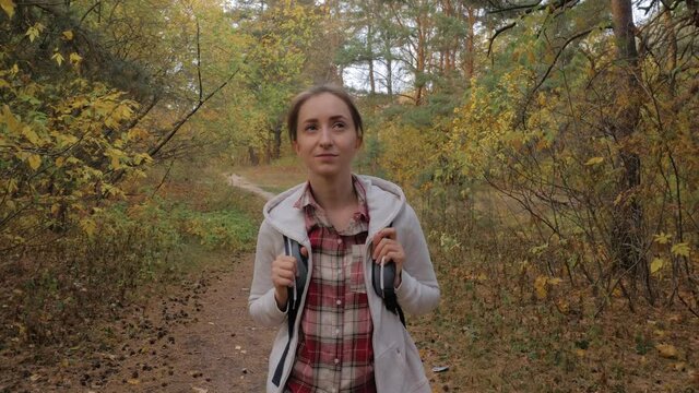 Front view of young positive woman with backpack walking in autumn park, forest and looking around - steadicam shot. Active outdoor lifestyle, leisure time, freedom and adventure concept