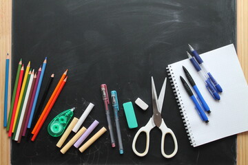 Top view of school items: notebook, pens, plasticine, crayons, erasers, scissors, pencils on the blackboard. Back to school, flat lay with copy space 