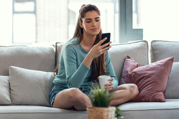 Pretty young smiling woman using mobile phone while drinking cup of coffee sitting on a couch at home.