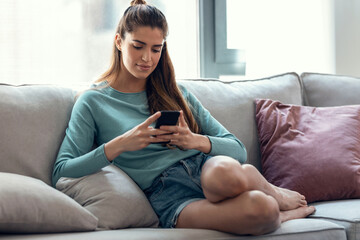 Obraz premium Pretty young smiling woman using mobile phone while taking a break sitting on a couch at home.