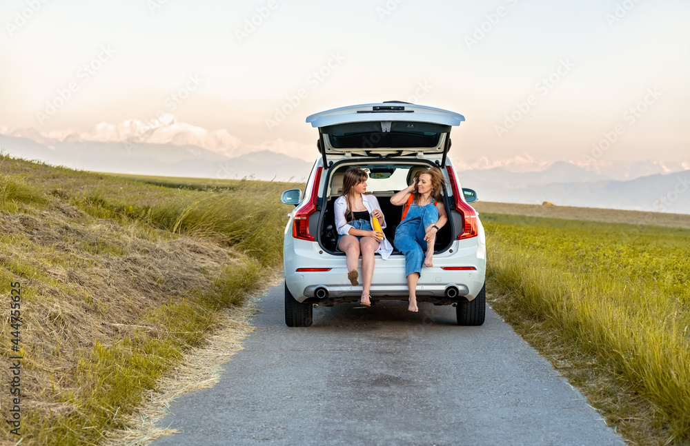 Wall mural female friends having fun enjoying their road trip in countryside sitting in open car boot having dr