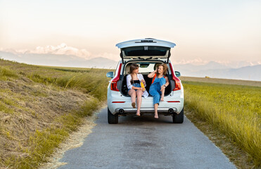 Female friends having fun enjoying their road trip in countryside sitting in open car boot having...