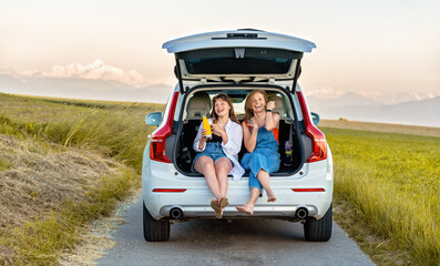 Mother and teenager daughter having fun enjoying their road trip in countryside sitting in open car boot having drink. Local traveling, summer holidays lifestyle.