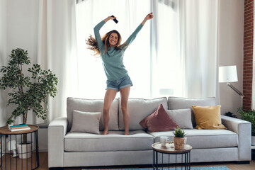 Beautiful woman listening to music with smartphone while dancing on the couch in the living room at home.
