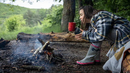 A girl on a hike sits near the fire and drinks a hot drink