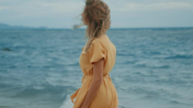 A slim blonde in a yellow dress walks along the beach and looks around