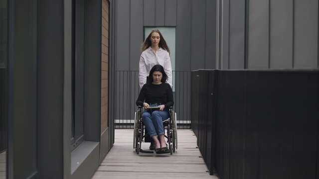 Wide shot portrait of young Caucasian woman pushing wheelchair with disabled colleague outdoors. Beautiful slim coworker taking care of paraplegic invalid. Support and inclusivity