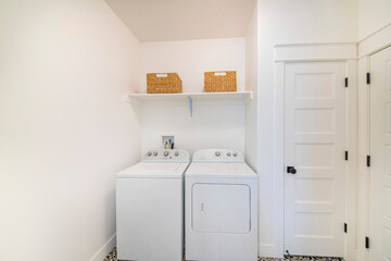 Small white laundry room interior with washer and dryer unit beside a narrow white door - obrazy, fototapety, plakaty