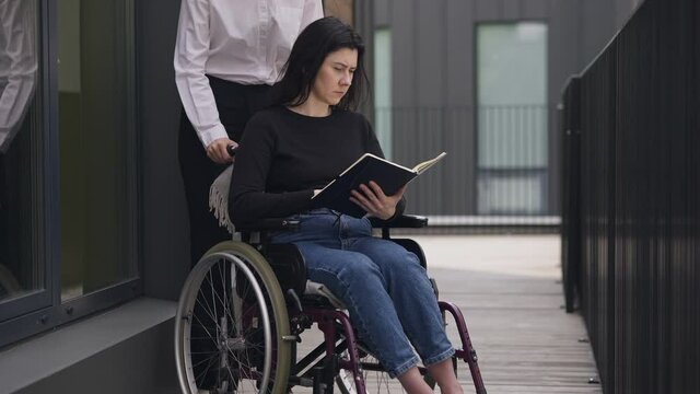 Absorbed concentrated disabled woman reading paperwork as unrecognizable assistant walking pushing wheelchair. Professional paraplegic Caucasian expert analyzing strategy