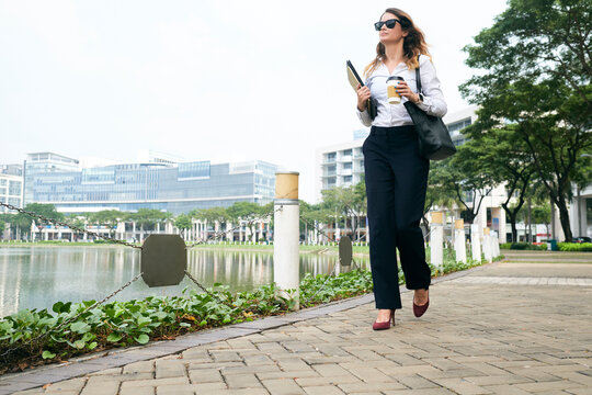 Confident female entrepreneur walking to office with cup of morning coffee and documents folder in hands