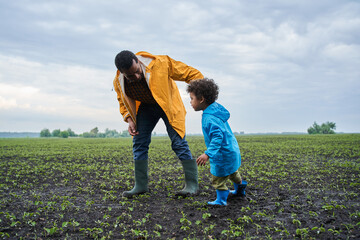 Father and son wearing raincoats and rubber boots exploring swampy field with green plants