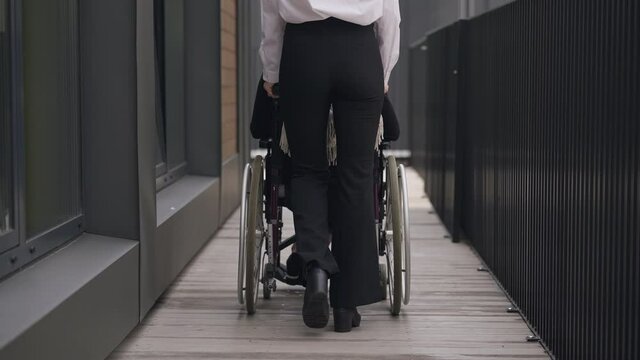 Back view of slim young Caucasian assistant pushing rolling wheelchair with handicapped CEO outdoors leaving. Wide shot of elegant woman taking care of disabled coworker. Business and inclusivity